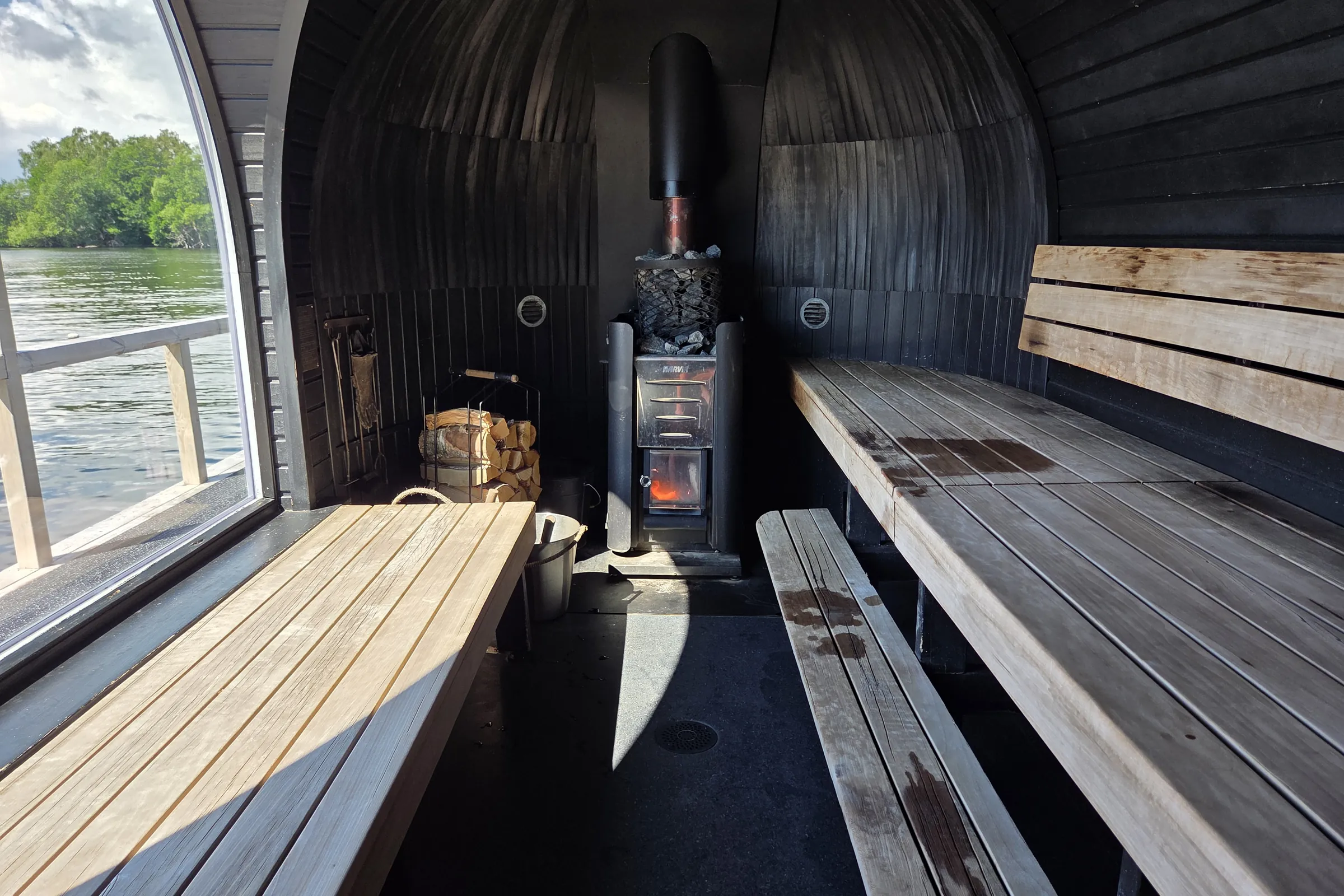 Inside the wood-fired sauna at Vinterviken Interior of the barrel sauna with black timber walls, tiered wooden benches along both sides, a wood-burning stove topped with sauna stones and a fire burning behind the glass, stacked firewood and a metal water bucket, with a tall window onto the water on the left.
