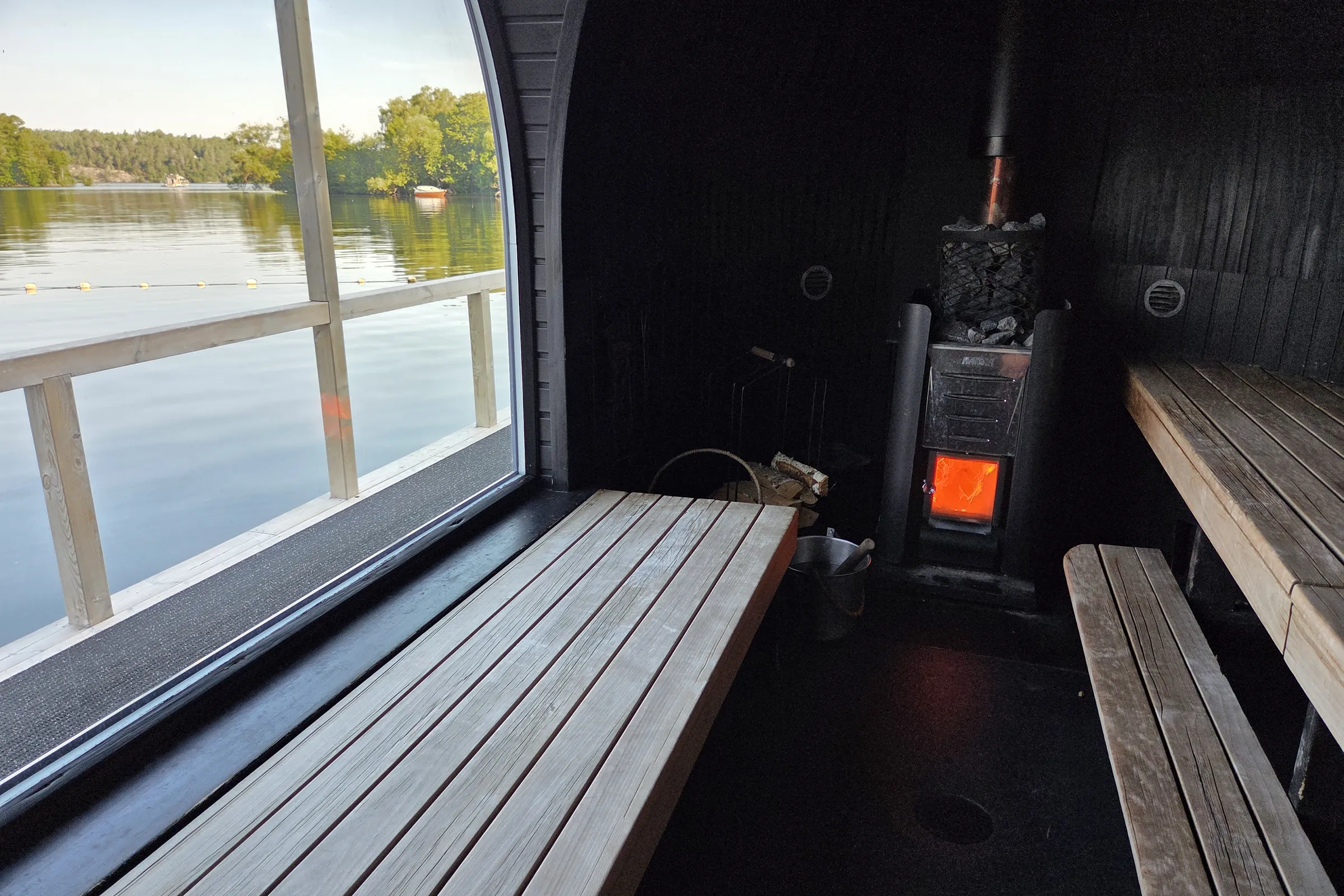 Bench with bay view inside the Vinterviken sauna Low view along a wooden sauna bench beside a large window overlooking Vinterviken bay, with a line of swim buoys on the water outside and the stove glowing orange in the dark interior on the right.