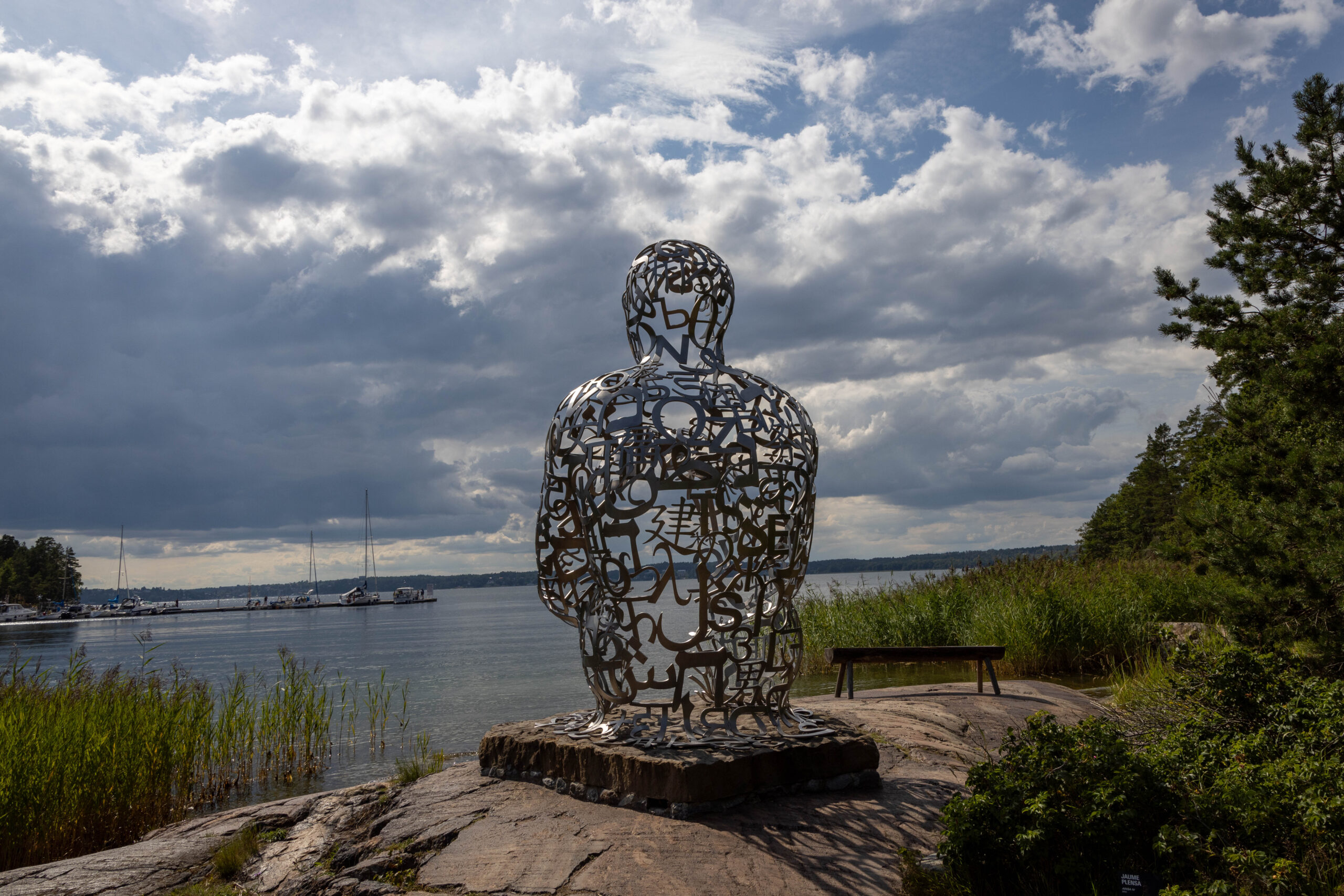 Outdoor art installation of a human shape sitting down looking out at the water. Statue at art museum Artipelag outside of Stockholm Sweden.