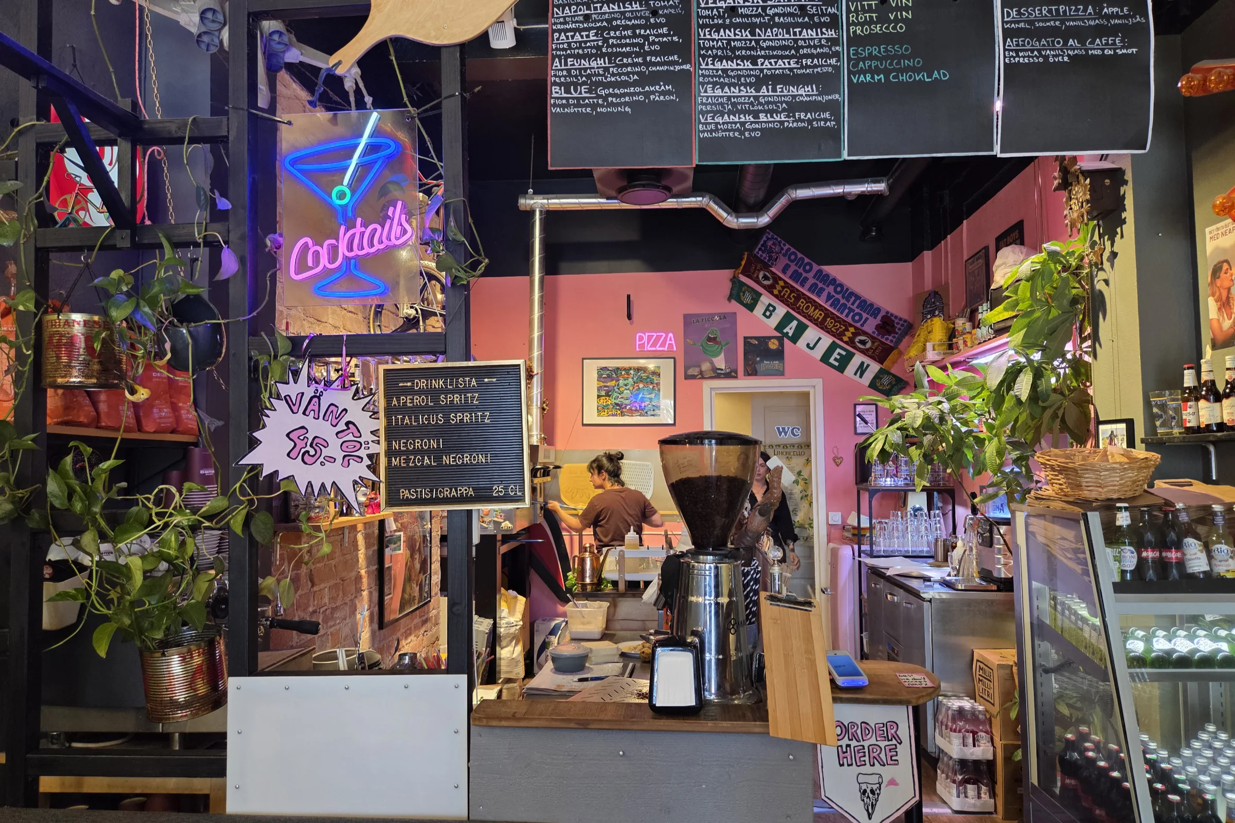 The colorful interior design at La Piccola Nonna in Stockholm. The desk where you place your order, and the pizza menu is written on a chalkboard above. There is a neon sign spelling cotails with a martini glass haning on the side and you can seee the kitchen in the background.