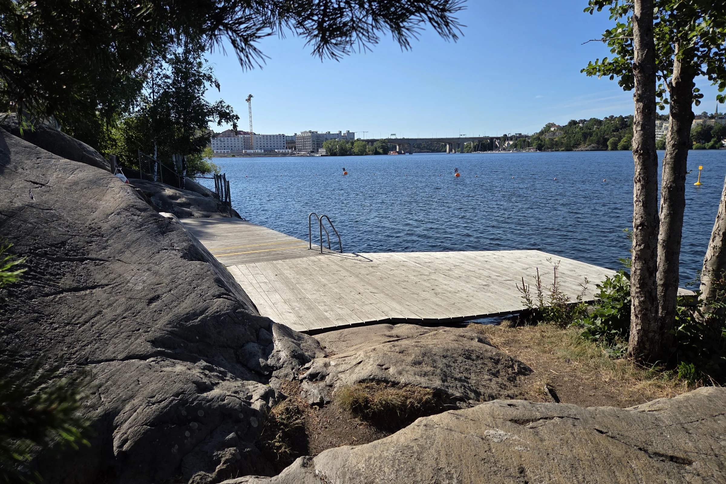 Fredhällsbadet bathing jetty on a sunny summer day, with some threes giving shades in the foreground and views towards the construction site of the new residential area at Lilla Essingen on the other side of the water.