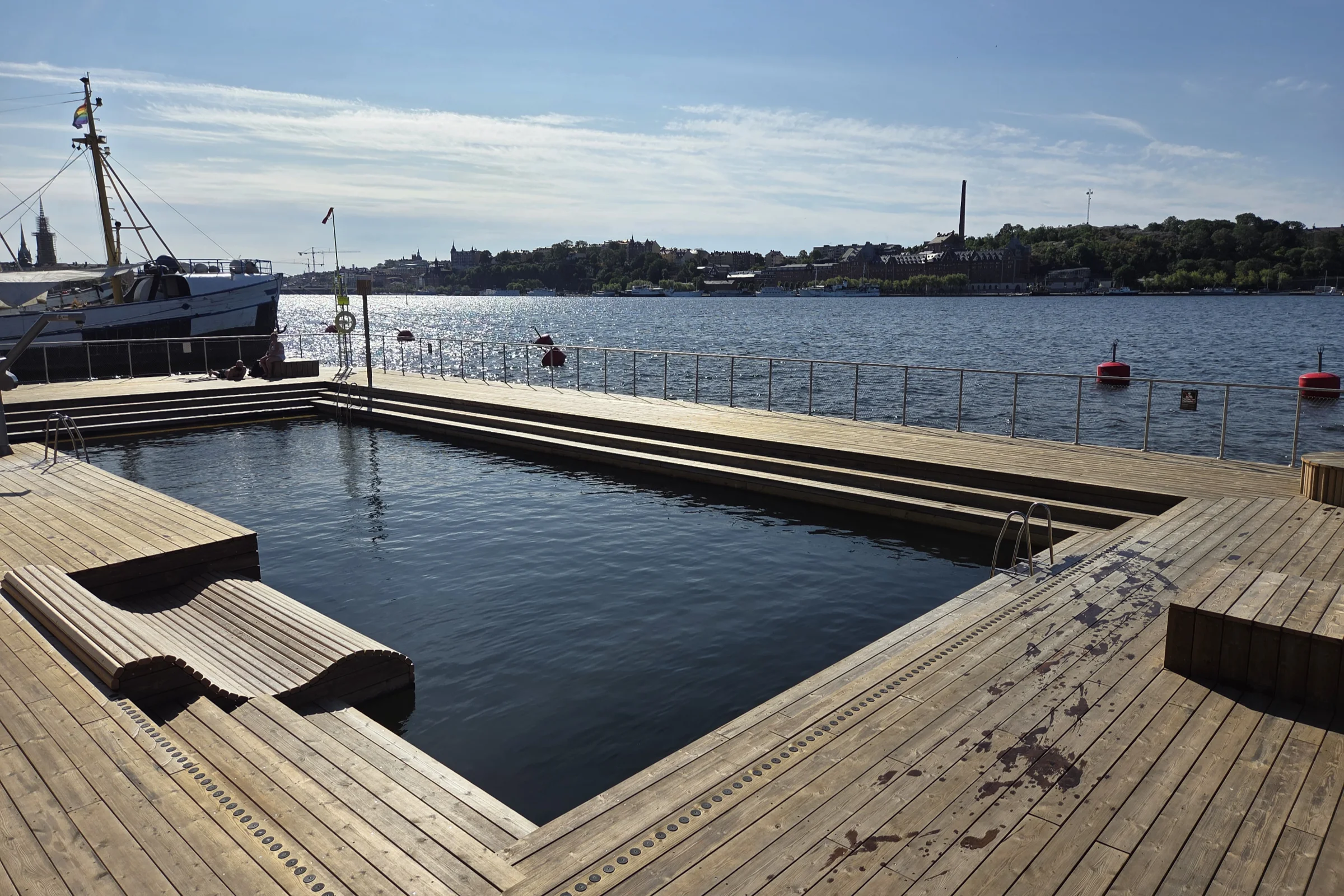 The harbour pool at Norr Mälarstrand in Stockholm on a sunny summer morning, next to the ships in the harbour and views of the Södermalm island across the water.