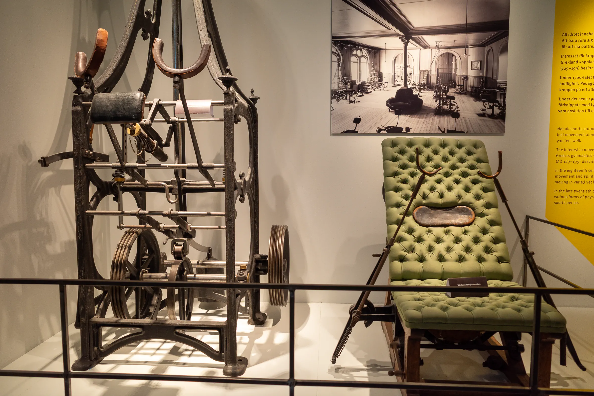 Antique mechanical exercise machines and a tufted green exam bench on display at Riksidrottsmuseet, with a historic gym photo behind