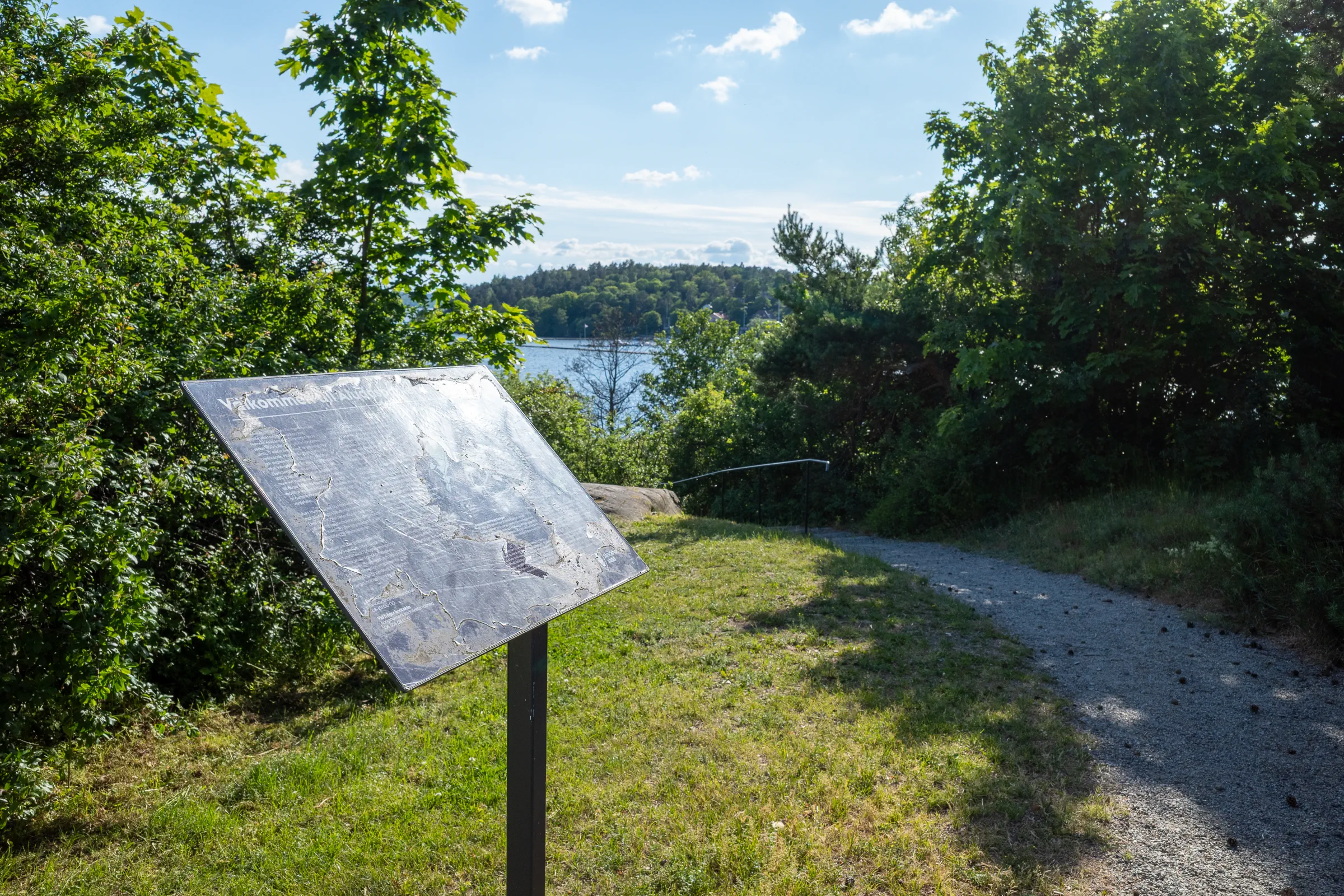 A worn information sign beside the gravel waterfront path on Stora Essingen, with Lake Mälaren visible through the trees