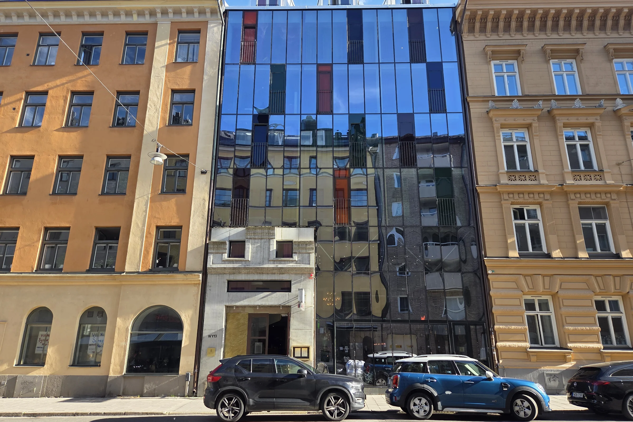 The exterior of hotel Hellstens Glashus in  Stockholm. The mirrored glass walls that mirrors the street in front of the hotel, and with a few cars parked in the front of the hotel.