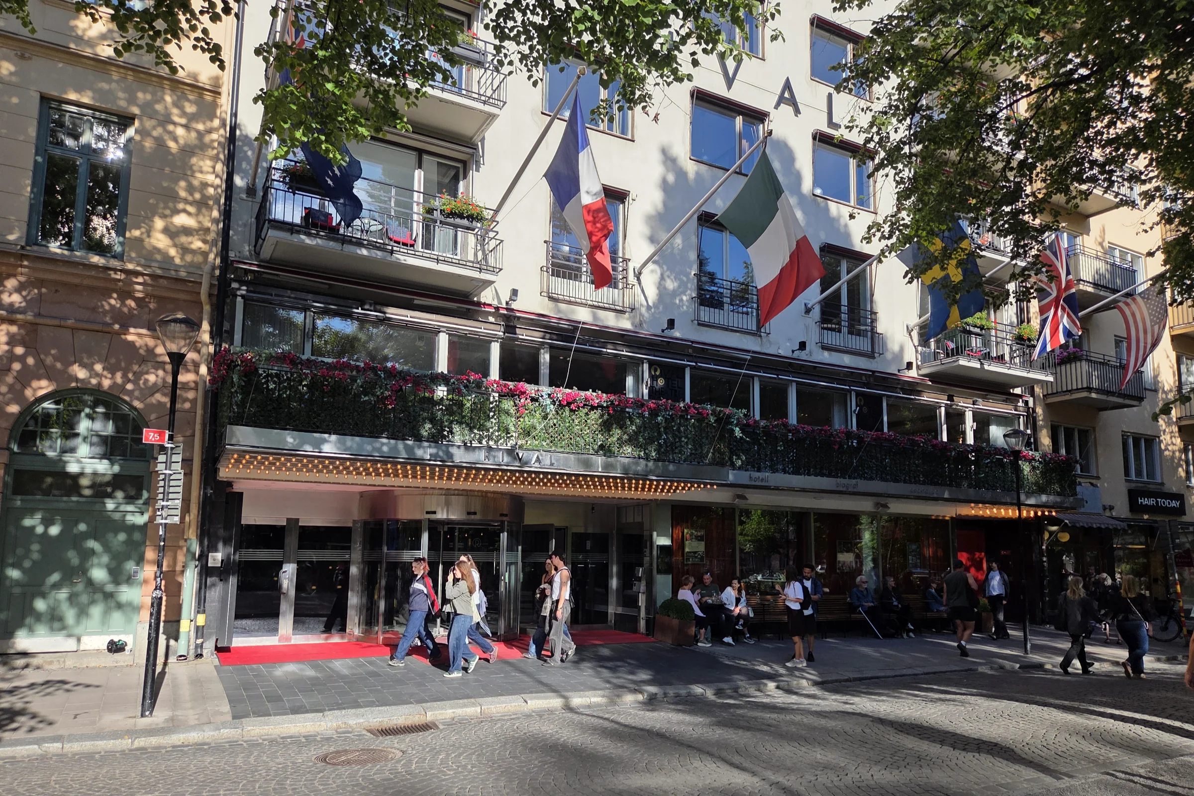 The exterior of Hotel Rival at Mariatorget in Stockholm on a sunny summer day. The French, Italian, Swedish, British and American flags are hanging above the entrance, people are walking past and a few visitors are sitting on the benches in front of the hotel. Red flowers are decorating the balcony a floor above the entrance floor.