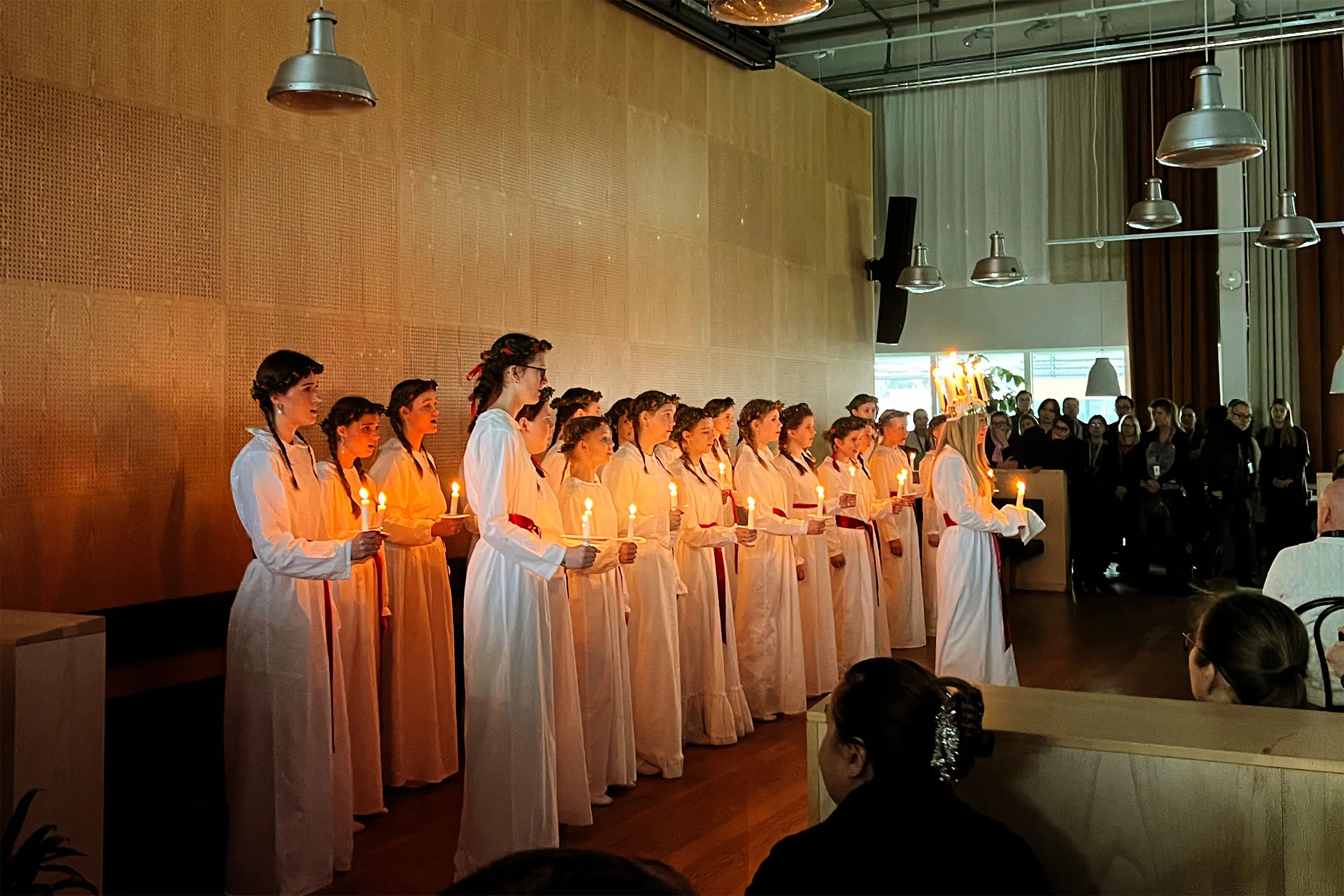 A traditional Swedish Lucia procession featuring a choir of white-robed singers holding candles and wearing crowns of lights, performing in an indoor setting.